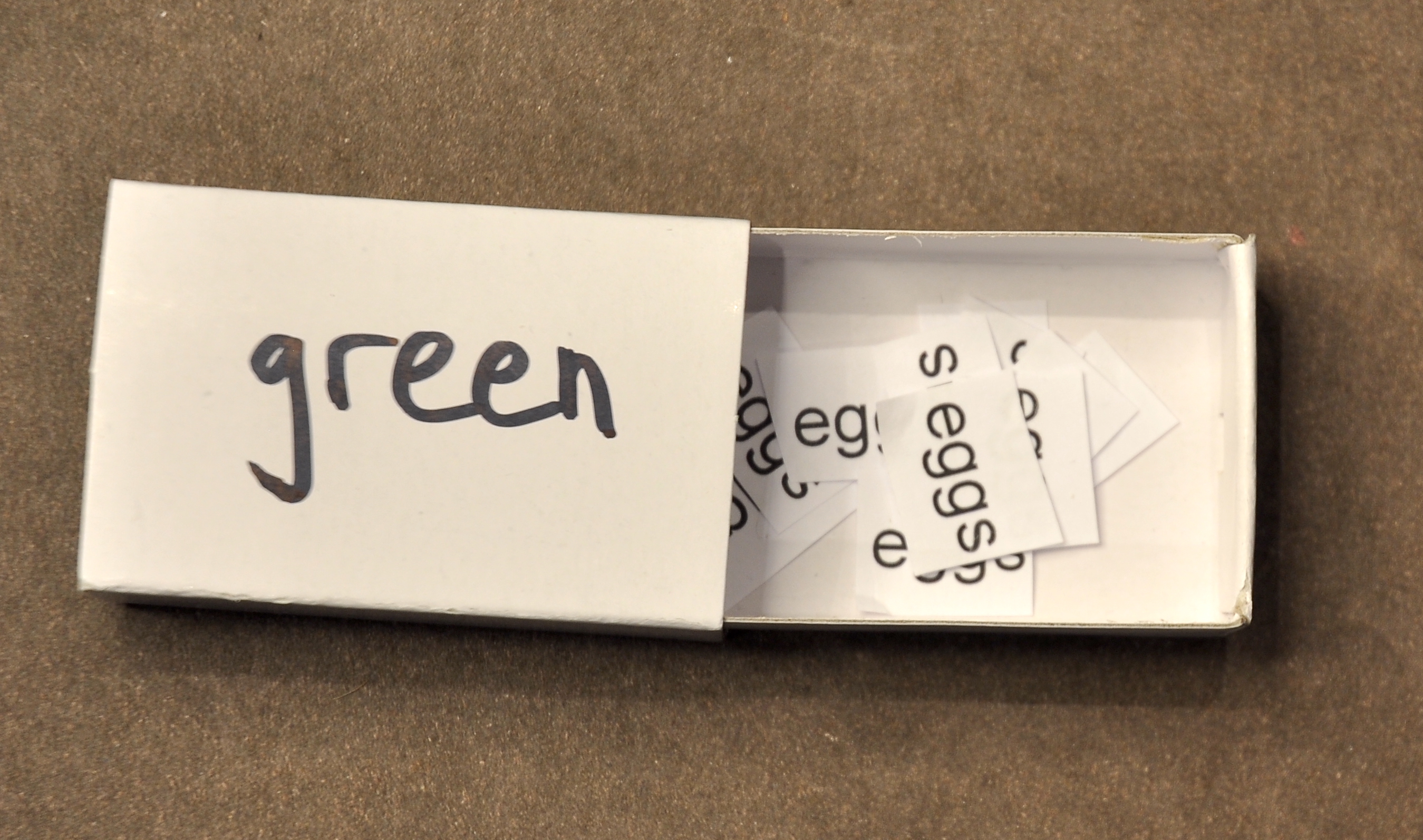 a matchbox with the word 'green' written on top, open to show the word eggs on multiple slips of paper